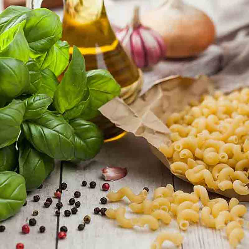 Basilikum, Maccheroni, Olivenöl, Knoblauch, Pfefferkörner auf Holztisch.