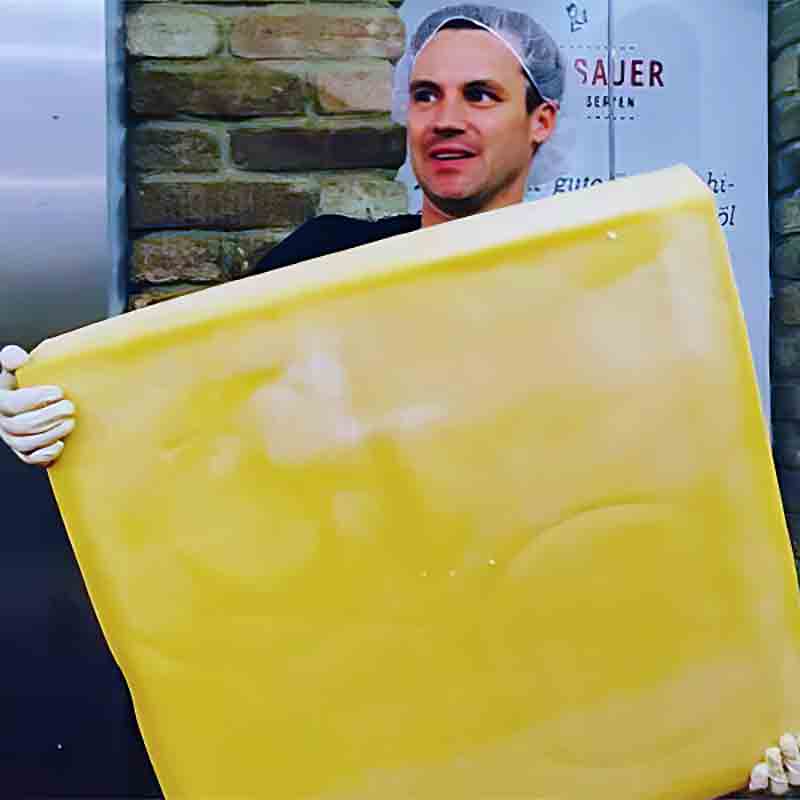 Stefan Grossauer trägt einen sehr großen Hartkäse in den Händen.