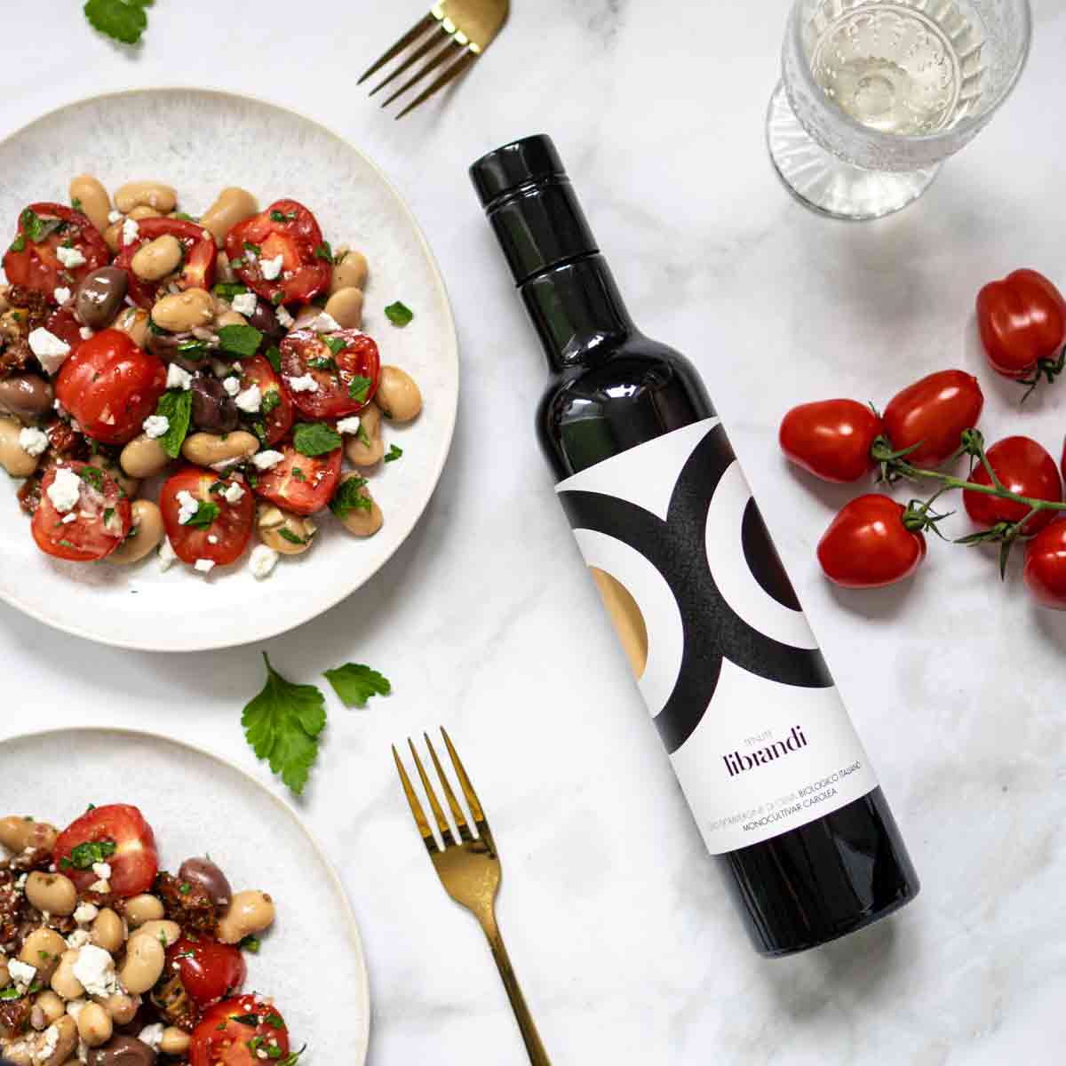 Olivenölflasche Librandi, Bohnensalat, Tomaten, Gabeln, Glas Wasser.