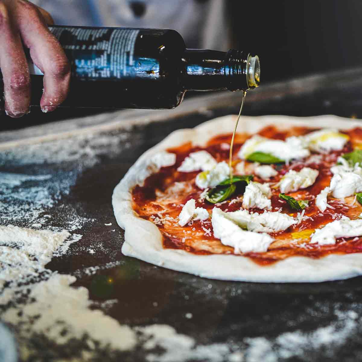 Hand gießt Olivenöl auf Pizza mit Tomaten und Käse.