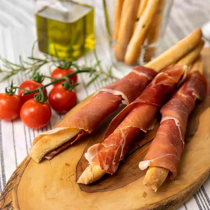 Grissini mit Schinken, Olivenöl, Tomaten auf Holzplatte.