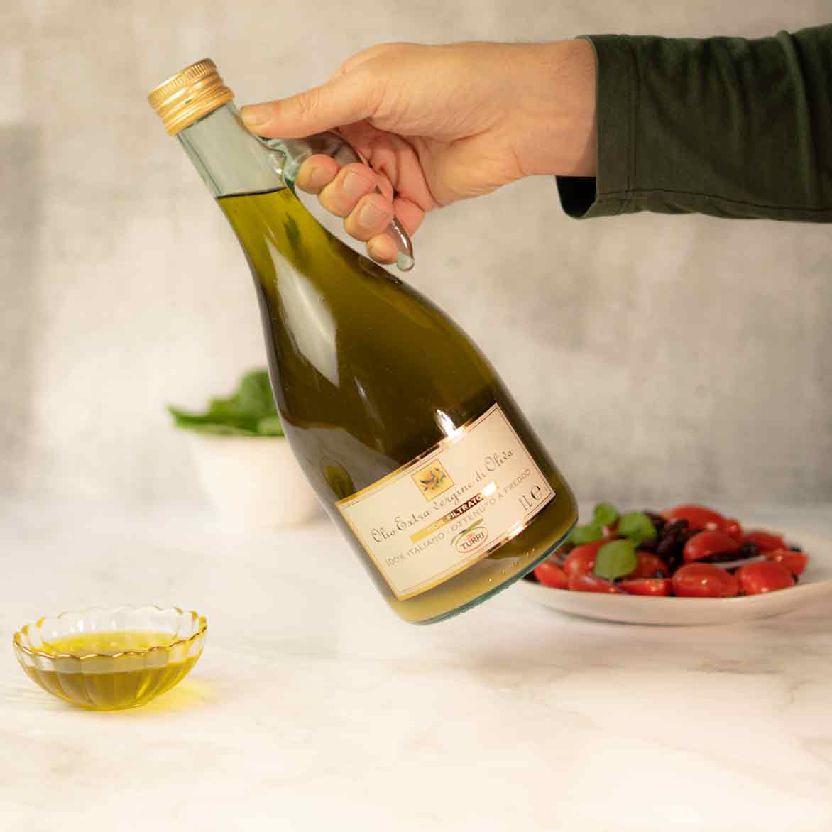 Hand hält Turri Olivenölflasche, Schale und Teller sichtbar.