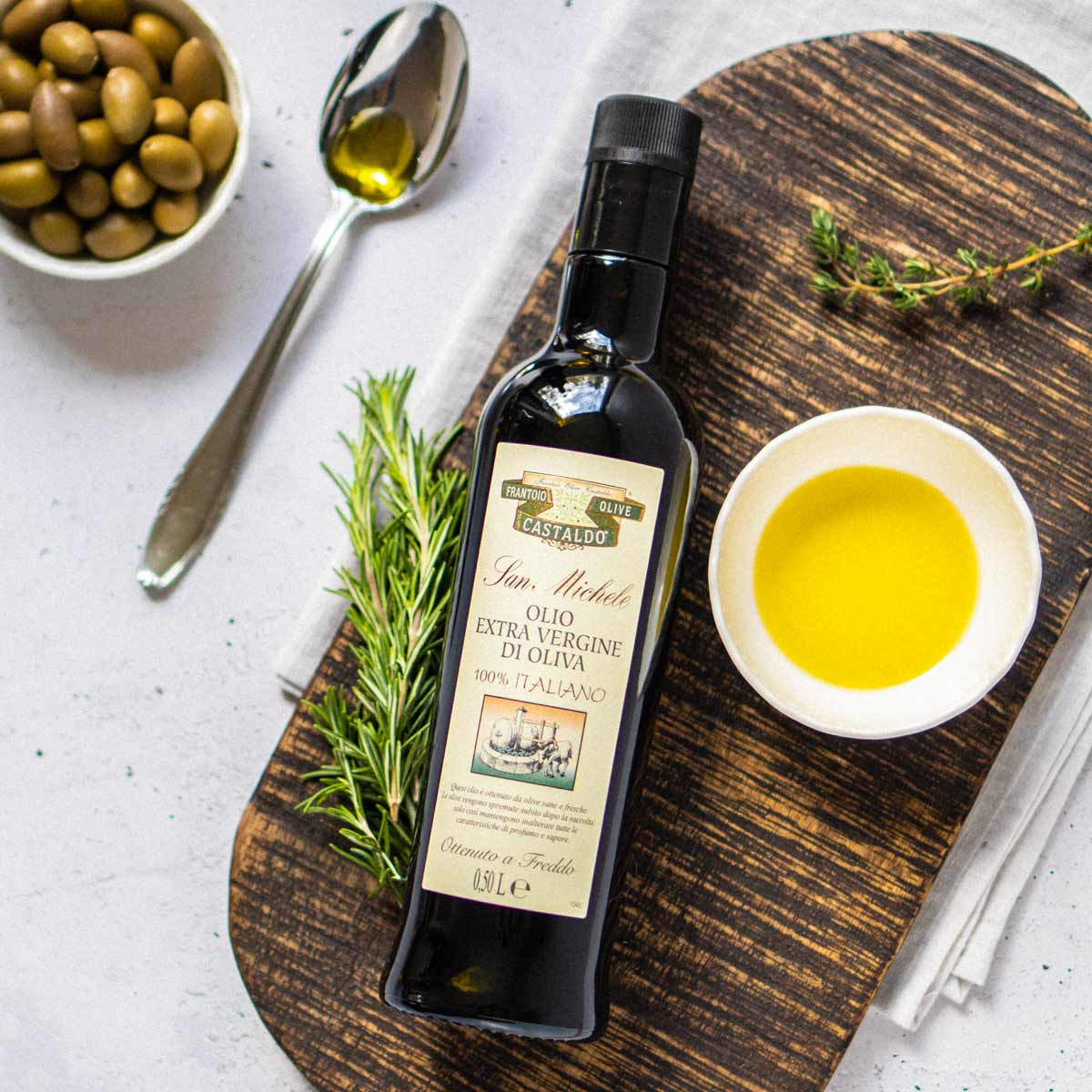 Olivenölflasche San Michele mit Oliven und Kräutern arrangiert.
